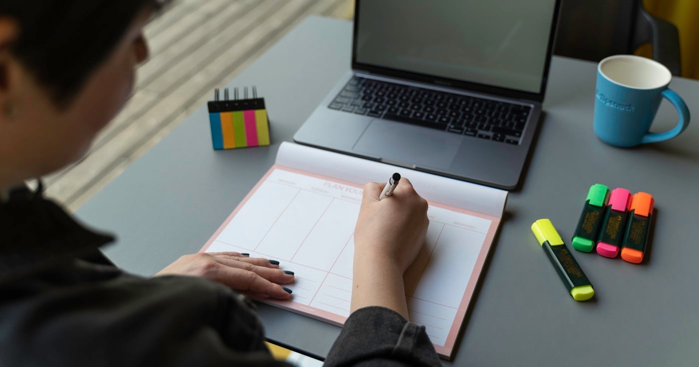 Person writing in a planner at a desk with sticky notes and laptop
